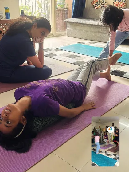 A group of young girls in a yoga class, one resting while others are in different poses. The atmosphere is relaxed and non-competitive.