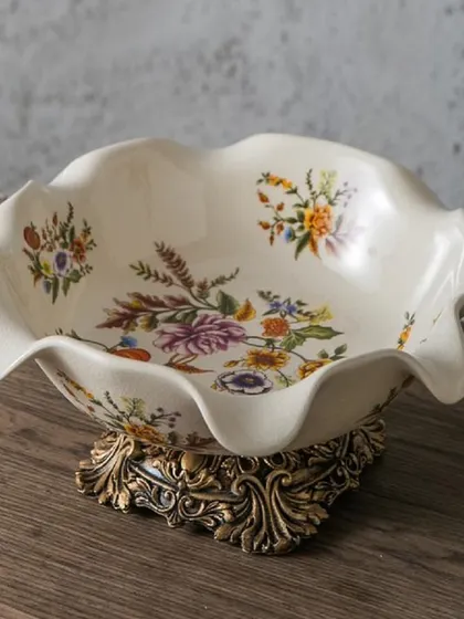 This decorative bowl is a perfect example of Victorian charm, with its scalloped edges, delicate floral paintings, and an ornate, antique-gold finished base. It's ideal for use as a fruit bowl or a standalone centerpiece.