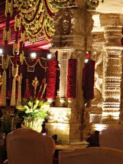 A close-up view of the intricately carved pillars and the vibrant floral arrangements. The pillars are wrapped in red flowers, contrasting beautifully with the stone-finish texture and the traditional garlands.