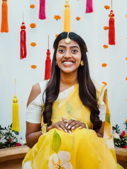 A portrait of the beautiful bride in her yellow saree, seated before the colorful tassel backdrop, looking radiant and happy.