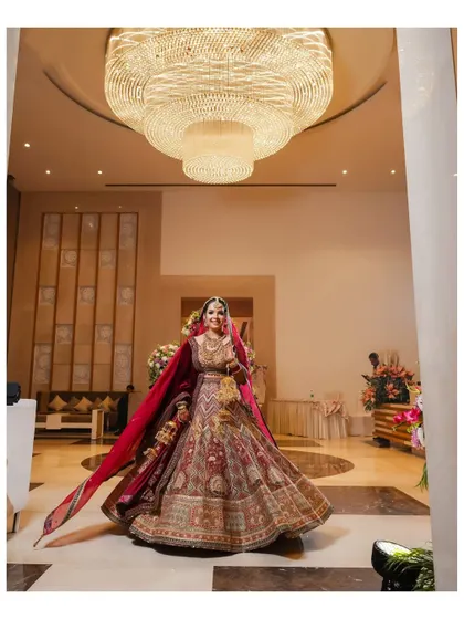 A grand, full-length portrait of the bride in the hotel lobby. The wide-angle shot captures her stunning lehenga and the opulent chandelier, creating a sense of scale and luxury.