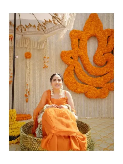 The bride, glowing in orange, sits for her Haldi ceremony against a backdrop of a large Ganesha made from marigold flowers.