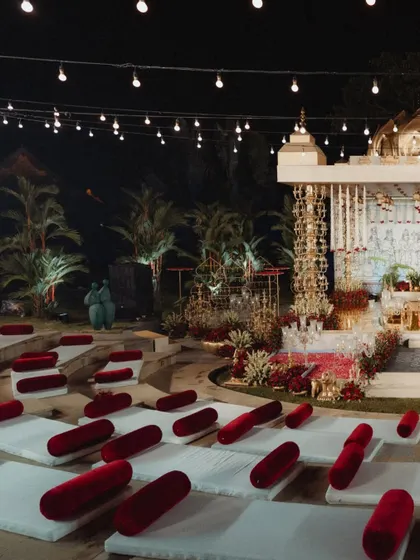 A side view of the amphitheater at night, showing the glowing mandap and the tiered seating dotted with red velvet cushions. The string lights in the background add to the magical, festive atmosphere.