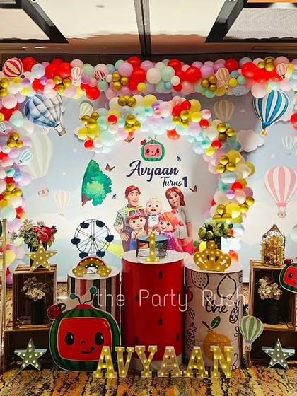 Avyaan's first birthday was a colorful Cocomelon celebration. The setup includes a large backdrop with the whole Cocomelon family, character cutouts, and a vibrant balloon arch with hot air balloon accents.