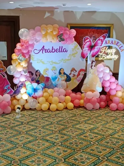 A Disney Princess themed birthday party with a custom backdrop. The setup features cutouts of Aurora and Belle, butterfly accents, and a pink and yellow balloon arch.