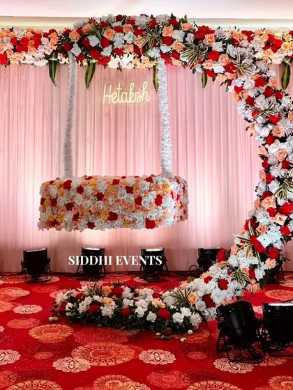 A whimsical floral swing, covered in red, peach, and white roses, suspended from a matching floral arch. A custom neon sign with the baby's name makes it perfect for a naming ceremony.
