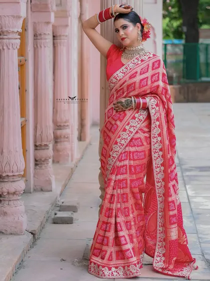 A full-length portrait against a pink architectural backdrop. The model's pose is confident and stylish, showcasing the full drape of the red saree.