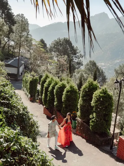 A high-angle shot of a couple walking down a beautiful, tree-lined path in the hills of Mussoorie. The vibrant colors of their outfits pop against the lush greenery.