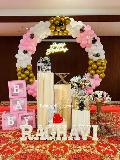 A circular balloon arch in pink, white, and gold for Raghavi's birthday. We added personalized touches like light-up marquee letters and "BABY" blocks to make the setup feel special.