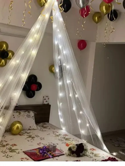 A cozy and romantic bed canopy decorated with fairy lights, with balloons on the ceiling and gifts waiting on the bed.