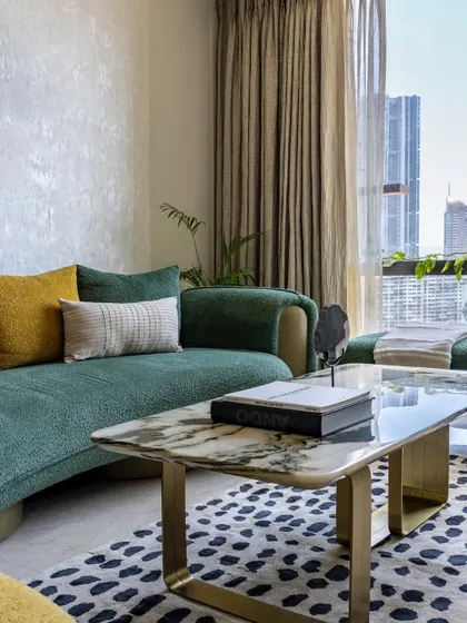 A close-up of the green curved sofa with textured fabric, paired with a marble coffee table and a bold patterned rug. The neutral walls allow the vibrant colors of the furnishings to stand out.