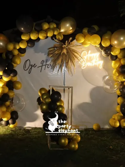 The main photo backdrop for the 40th birthday featured a white panel with a neon 'Happy Birthday' sign, framed by a dramatic black and gold balloon arch and accented with dried floral arrangements.