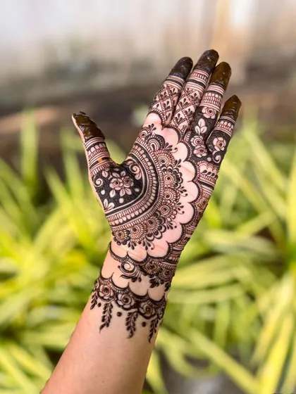 A single hand showcasing a modern henna design. The pattern features a large floral element with a scalloped border, combining fine lines with bold, filled sections for a striking look.