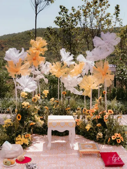 The main seating for the Haldi ceremony, set against a backdrop of giant, ethereal paper flowers and a field of real sunflowers, creating a beautiful and artistic focal point.