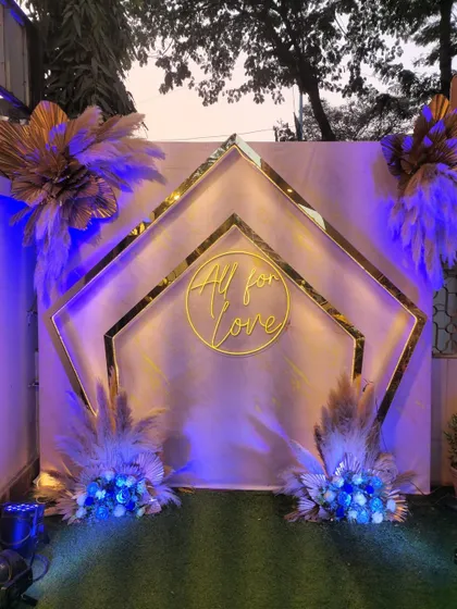 A modern bohemian photo booth with a blue-lit theme. The geometric gold frames, pampas grass, and "All for Love" neon sign create a stylish and contemporary look.
