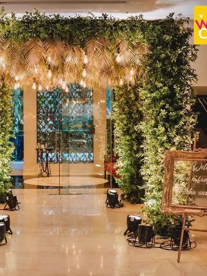 Welcome to the party! For this Sangeet night, I created a dramatic tropical entrance with dense foliage and gold-painted palm leaves, lit by warm Edison bulbs to set an energetic and glamorous mood.