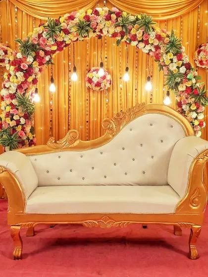 A classic and regal stage backdrop featuring a gold and white ornate sofa. The floral arch and hanging flower balls against a golden curtain create a traditional and festive look suitable for any ceremony.
