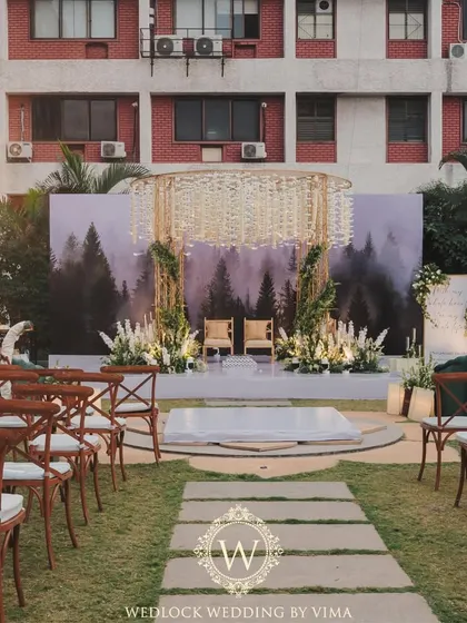A wide view of a minimalist wedding setup. The clean white aisle, lush green lawn, and a simple, elegant mandap against a forest backdrop created a serene and sophisticated atmosphere.