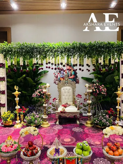 A complete Seemantham setup featuring a lush green backdrop with hanging floral garlands and a portrait of Krishna. The foreground is filled with beautifully arranged fruits and traditional offerings.