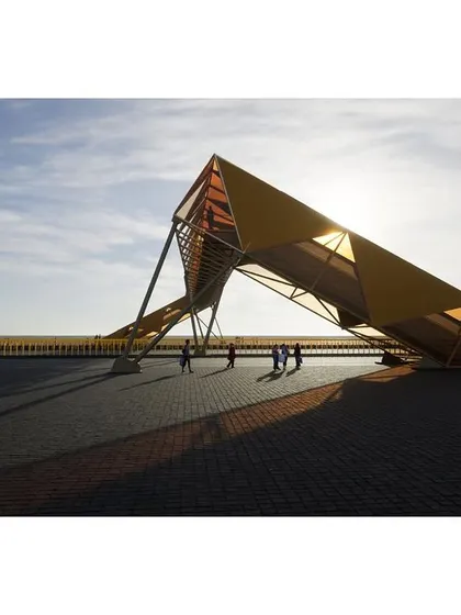 The entrance to the footbridge is designed to be an inviting public space. The structure's form creates dramatic shadows on the paved ground, enhancing the sense of arrival.