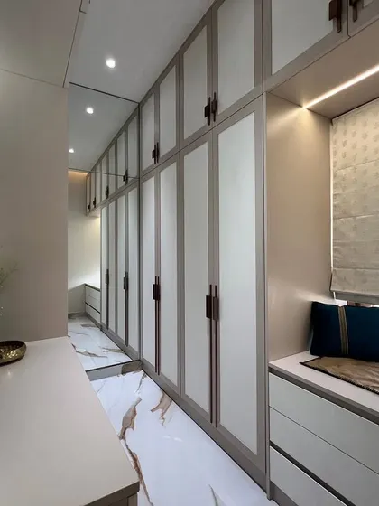 A spacious walk-in closet with floor-to-ceiling wardrobes in a neutral finish. The design includes a dressing table with a large mirror and a comfortable window seat with built-in storage.