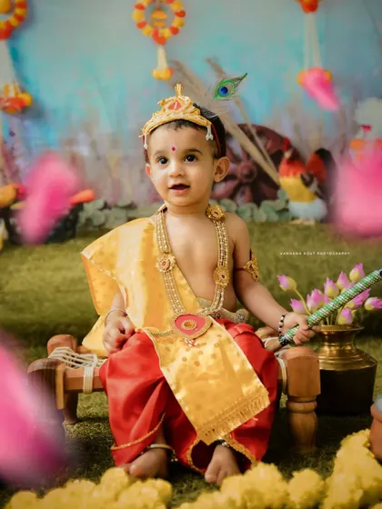 This little boy makes an adorable Krishna, dressed in red and yellow with a flute in hand. The festive backdrop is filled with details that celebrate the spirit of Janmashtami.