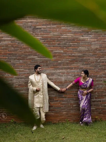 The couple holds hands, dressed in beautiful traditional attire for their engagement, with the groom in a classic sherwani and the bride in a vibrant purple saree.