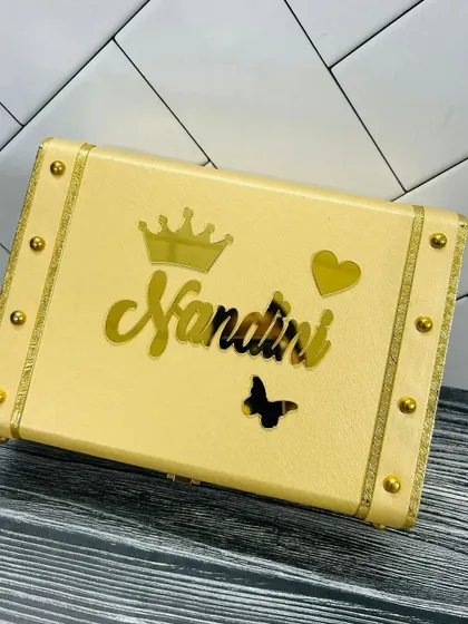 A cream-colored leatherette trunk box, customized with the name "Nandini" in gold acrylic, offering a sophisticated look.