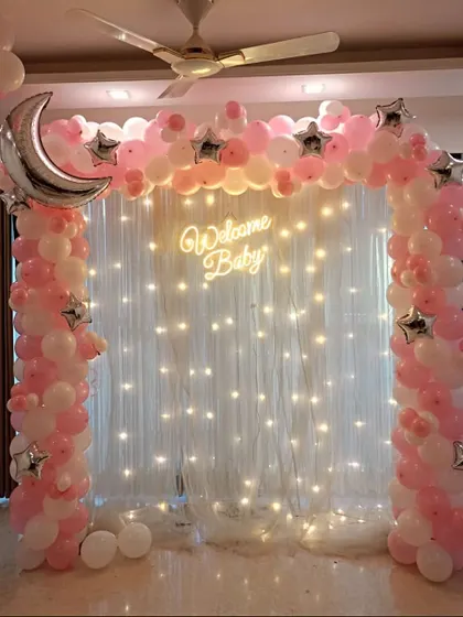 An elegant "Welcome Baby" setup with a fairy light backdrop. The pink and white balloon arch is accented with a crescent moon and star foil balloons.