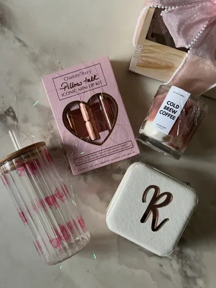 A flat lay of some of the luxury items I can include in a bridesmaid hamper, such as a Charlotte Tilbury mini lip kit, a personalized jewelry box, and a cold brew coffee candle.