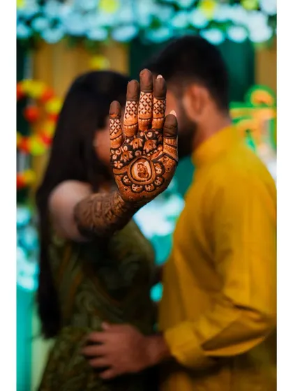 A romantic and creative mehendi photo, with the bride's henna-covered hand in the foreground and the couple sharing a quiet moment in the background.
