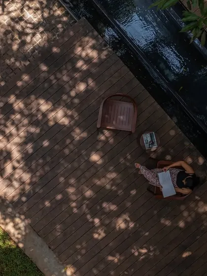 An aerial view of a resident enjoying the wooden deck at the 'Mango Tree Yard' home. This space was intentionally designed under the canopy of existing mango trees to create a perfect, shaded spot for reading and relaxing.