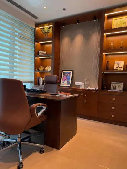 A wide-angle view of a sophisticated office cabin we furnished. This space includes a large wooden executive desk, ergonomic leather chairs, and a custom full-wall shelving unit with integrated backlighting for displaying awards and decor.