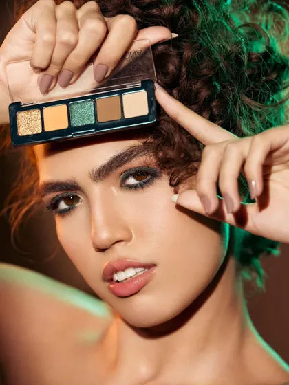 An inventive and playful pose where the model frames her eye with a Natasha Denona eyeshadow palette. The green and orange lighting complements the eyeshadow colors, creating a cohesive and artistic look. This shot is all about creative expression and the fun of makeup.