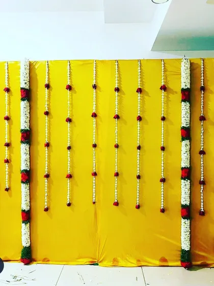 This backdrop uses a yellow cloth as a base, decorated with vertical garlands of jasmine and tuberose (rajnigandha), accented with red flowers. It's a clean and traditional floral decoration.