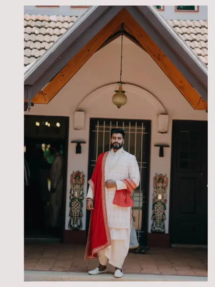 A striking portrait of the groom in his traditional white and red attire, standing confidently at the entrance of the venue.