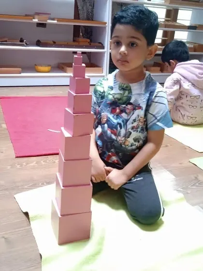 A child proudly sits with his completed Pink Tower. This sensorial material teaches size discrimination and prepares the child for mathematical concepts of cubing and the decimal system.