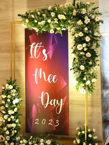 A closer view of the "It's Mee Day" photo backdrop, showcasing the vibrant, custom-printed banner and the lush white floral arrangements. We ensure every detail is sharp and perfectly executed for your event.