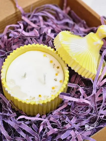 A close-up of a vibrant yellow candle in a fluted jar with a matching lid. The soy wax is decorated with gold stars and crystals, making the unboxing experience truly special.