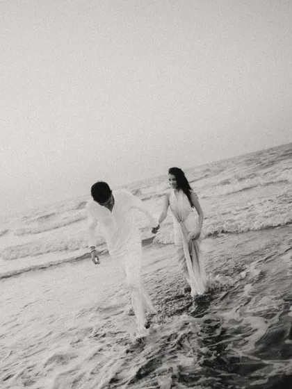 A candid black and white shot of the couple walking through the waves in Muscat. The image has a beautiful, grainy texture, capturing a spontaneous and joyful moment in a timeless style.