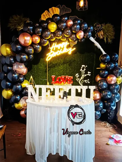 A chic and modern birthday setup with a black, gold, and rose gold balloon arch against a green hedge wall.