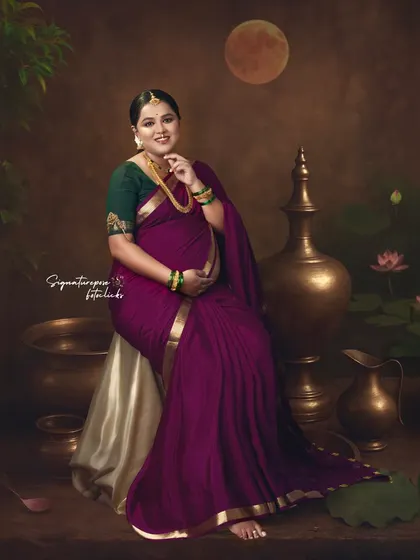 A seated portrait in a rich magenta saree, set against a painted backdrop with traditional brass vessels.