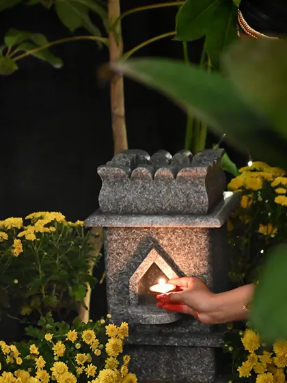 A hand places a diya into the niche of our granite Tulasi Brindavan, illuminating the sacred space amidst a bed of yellow flowers.