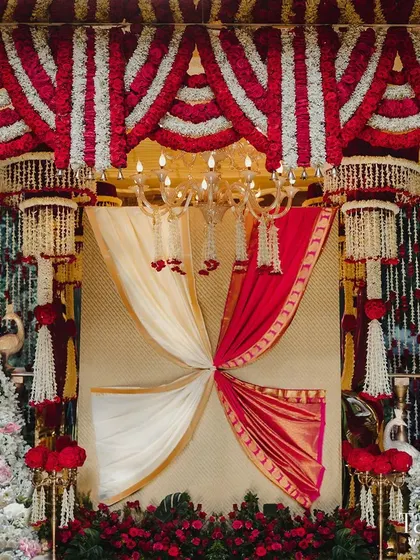 This backdrop is a tribute to South Indian tradition, featuring a beautiful drape of red and white fabric reminiscent of a saree and dhoti. It is framed by lush floral arrangements, creating a look that is both culturally rich and visually stunning.