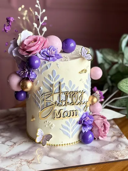 Delicate lilac and purple hues adorn this elegant cake for a mother's birthday. The design features beautiful stenciling, sugar flowers, and decorative spheres for a sophisticated look.