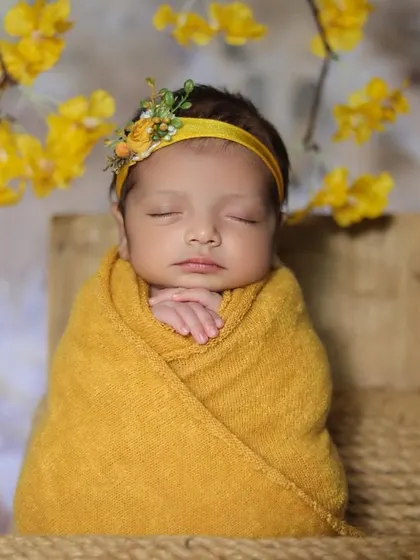 A sweet baby wrapped in a mustard yellow swaddle, sitting upright in a tiny jute chair. The delicate yellow blossoms in the background tie the whole look together.