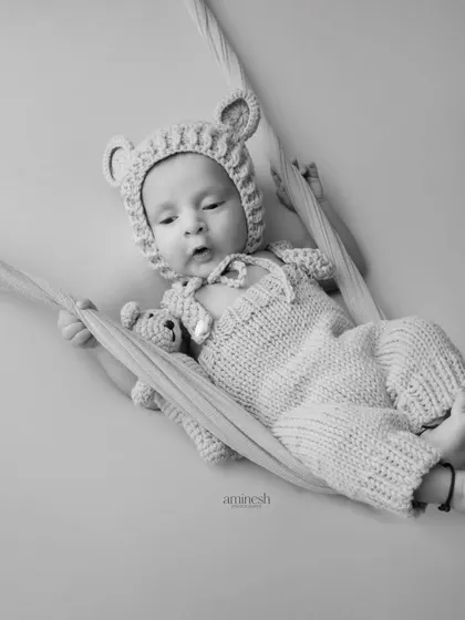 A timeless black and white portrait of a 20-day-old baby in a knitted bear outfit, holding a tiny teddy bear. The baby rests in a soft sling, creating a feeling of comfort and security.