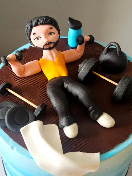 A close-up of the gym-themed cake topper. This view shows the details of the edible figurine lifting dumbbells, surrounded by other miniature workout equipment.