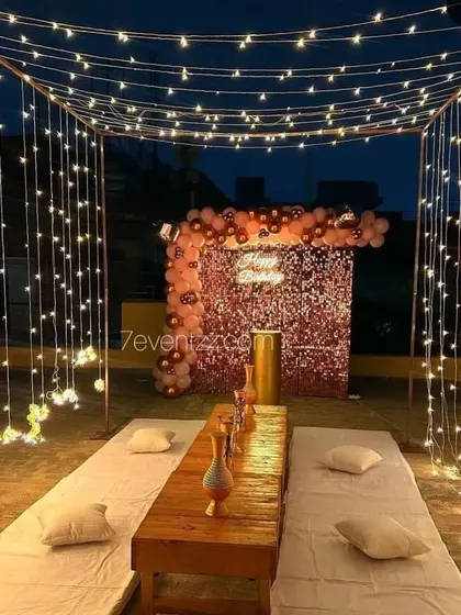 An enchanting rooftop dinner setup under a canopy of fairy lights, with a rose gold balloon backdrop.