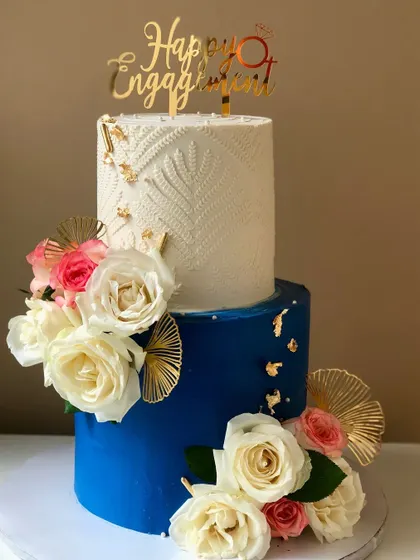 A stunning two-tiered engagement cake in royal blue and white, adorned with fresh roses and gold accents.